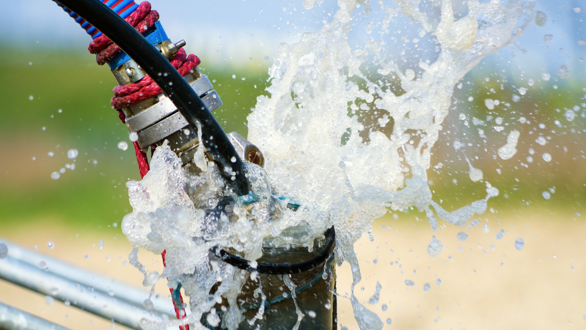 A close-up of a submersible well pump pipe spraying water forcefully, with splashes and hoses visible.