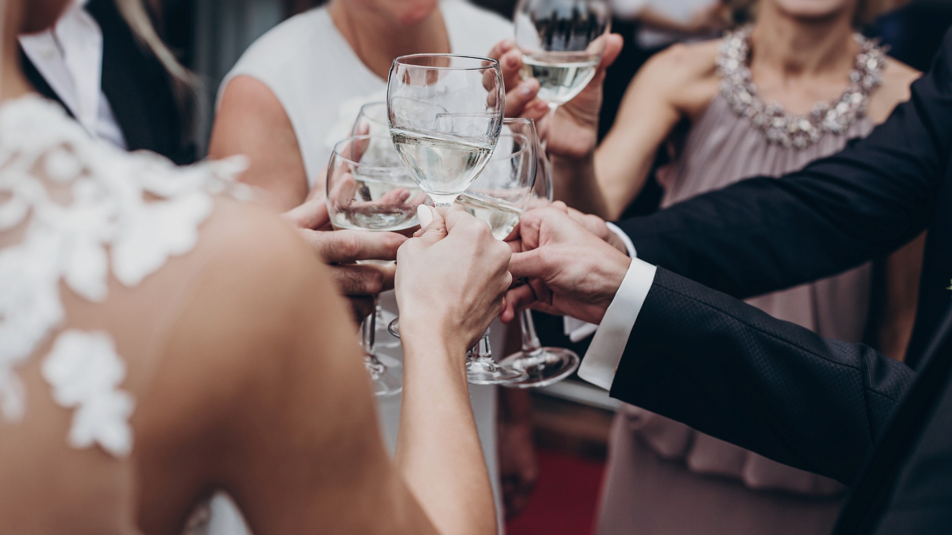 Six guests attending a wedding standing in a circle. All six guests toast with glasses of white wine.