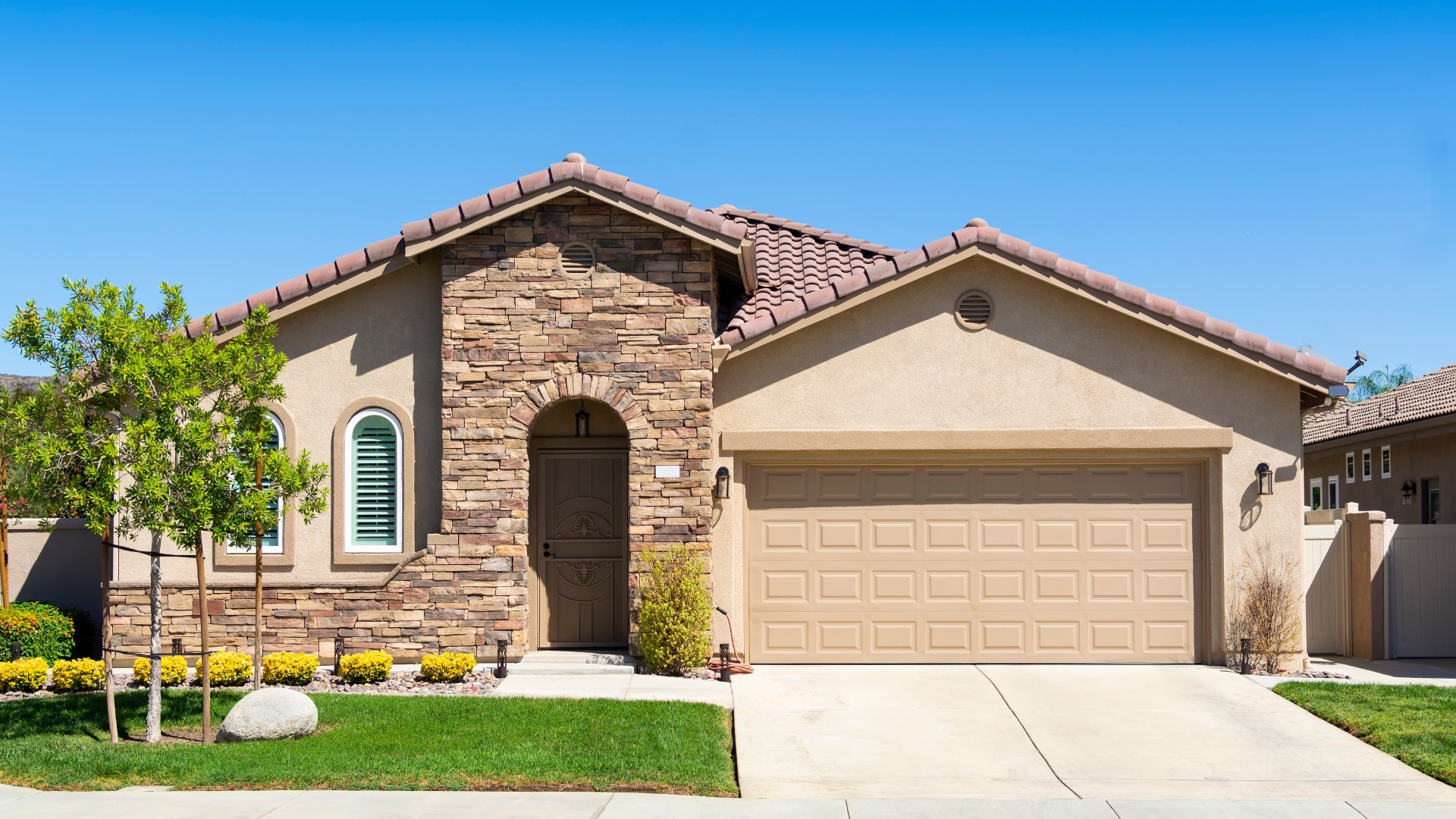 A beige home with a small concrete driveway installed in the front. The home has a clean lawn with four thin trees.