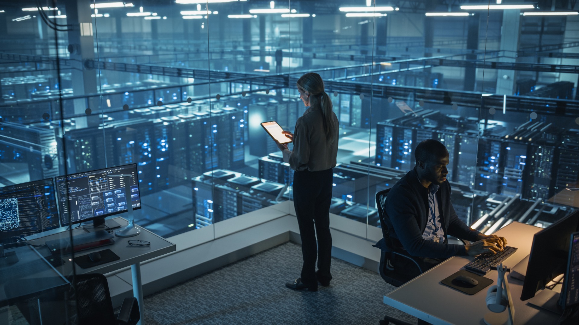 Two employees inside a data center. One employee looks at a tablet while another uses a desktop computer.
