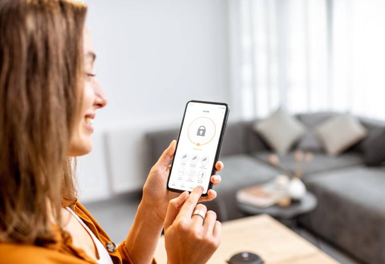 A young woman standing in her home, focused on her smartphone displaying a security application interface.