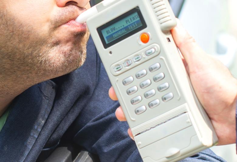 A close-up of a hand holding a modern breathalyzer with a digital screen and multiple buttons close to a man’s mouth.