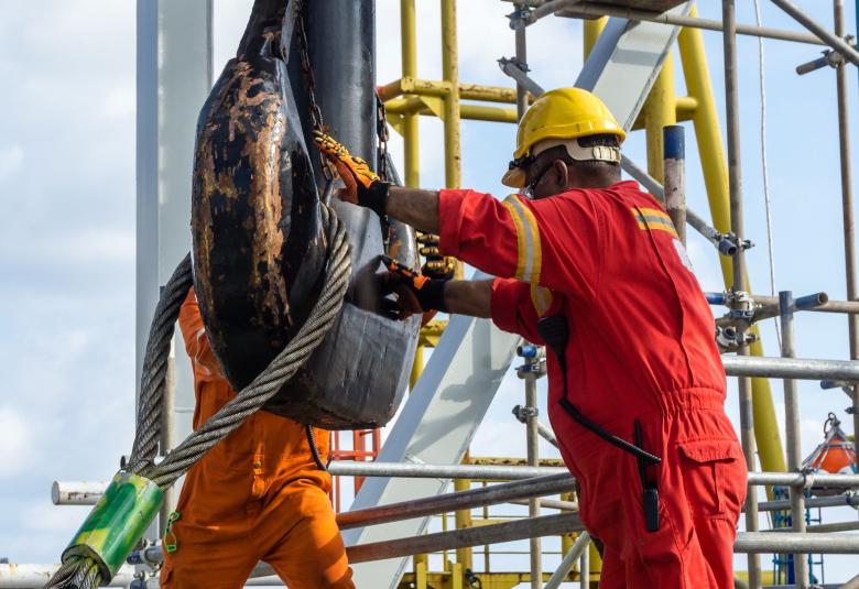 Two workers wearing orange and red jumpsuits attach a wire rope loop to a giant crane hook on a job site.