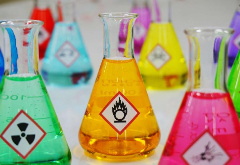 Multiple Erlenmeyer flasks filled with colorful chemicals. Each flask has a different hazardous warning symbol on the front.