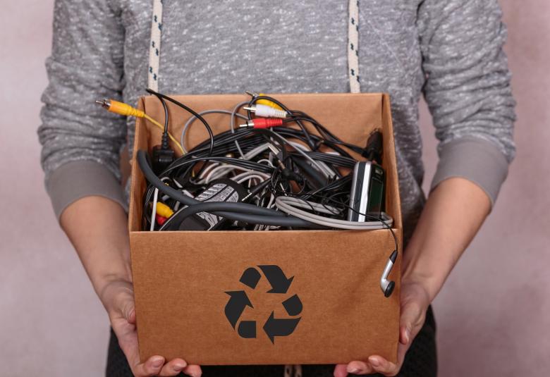 A close-up of female hands holding a cardboard box with a recycle symbol, filled with old digital devices.