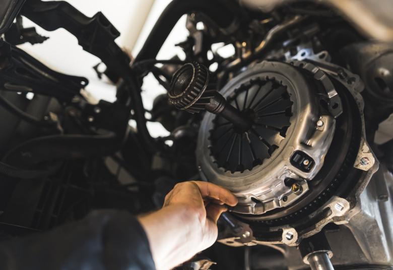 A close-up of a mechanic's hand reaching into the undercarriage of a lifted vehicle to touch the exposed clutch.