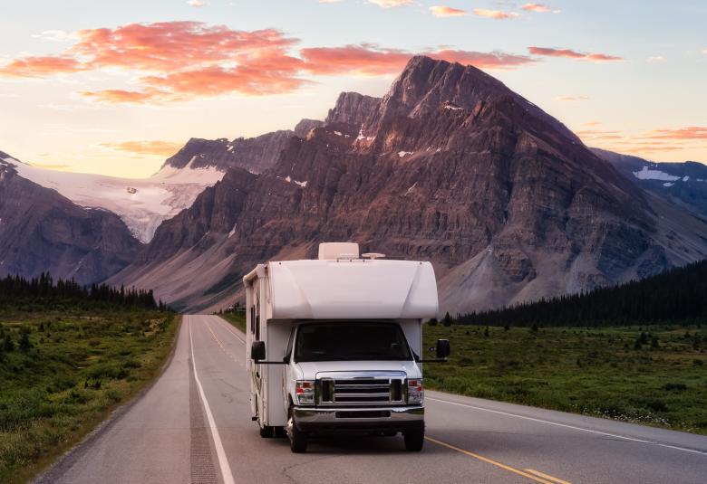 A white RV driving down an open road with a snowcapped mountain range and a sunset in the background.
