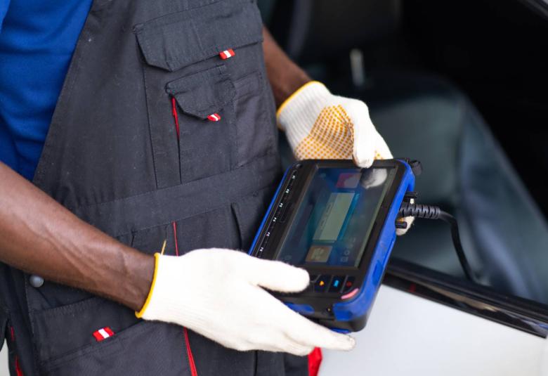 A mechanic in overalls and gloves is holding a device with a screen to read data from a car's engine control unit.