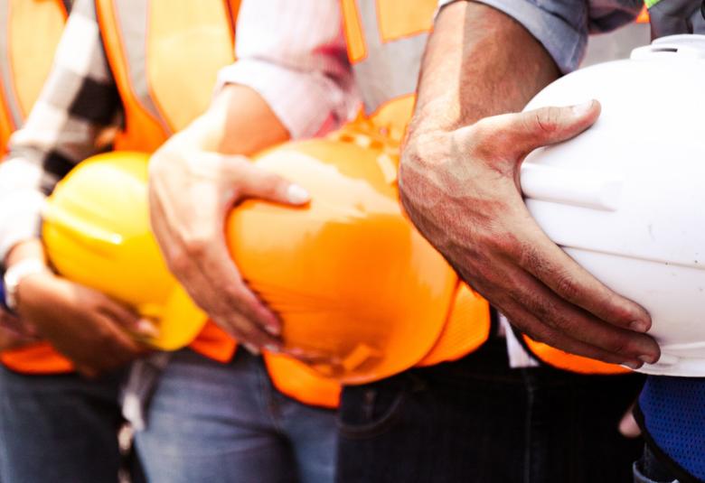 A close-up of a group of workers in a line, all wearing high-visibility safety vests and holding hard hats.