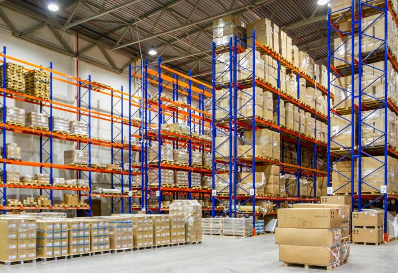 The interior of a large warehouse with an extensive pallet racking system. The pallets are filled with brown cardboard boxes.