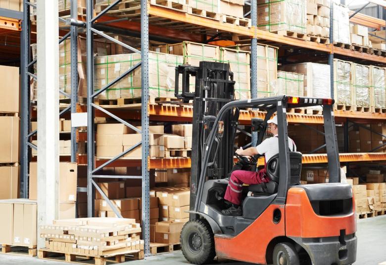 A warehouse employee wearing a white hard hat, maroon pants, and white shirt drives an orange and black forklift.