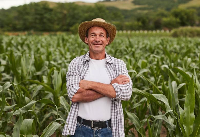 A happy farmer stands in front of his corn crop with his arms folded across his chest and a smile on his face.