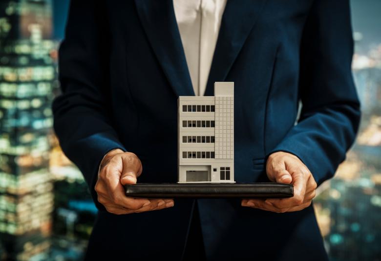 A businessperson holds the model of a commercial building on a flat surface while standing in front of a cityscape.