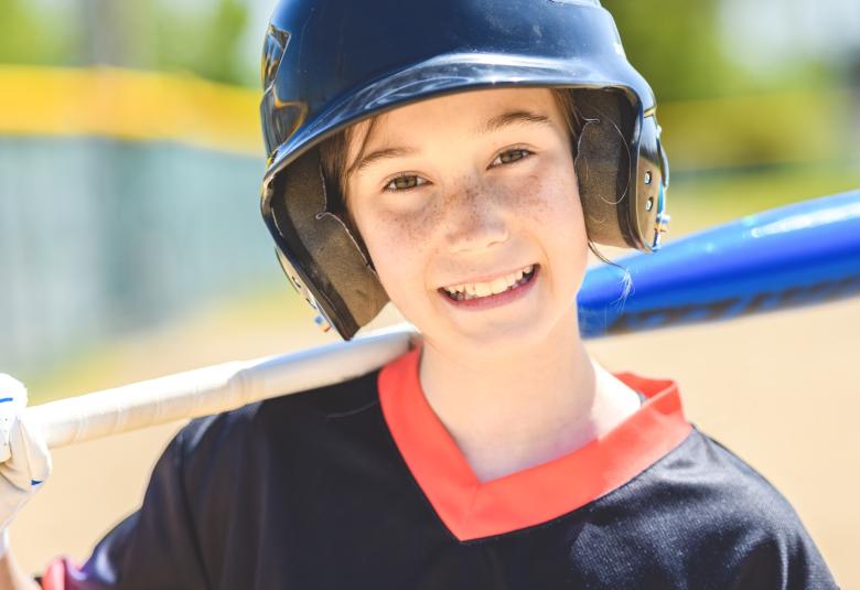 A close-up view shows a child wearing a black safety helmet and a black shirt. The child holds a blue bat over their shoulder.