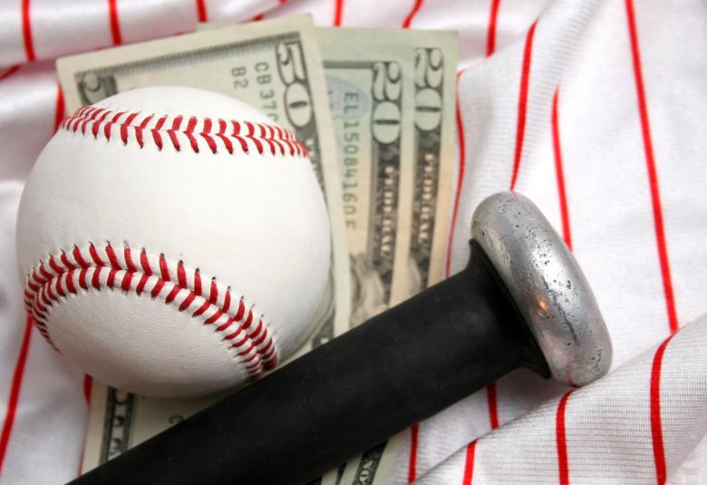 A close-up of a baseball and the knob of a baseball bat resting on three dollar bills and a white baseball jersey.