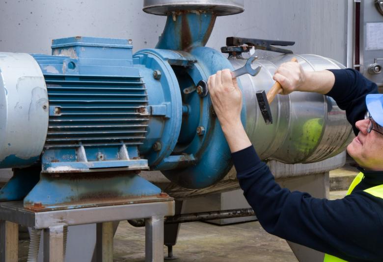 A man wearing a blue hard hat, a black long-sleeve shirt, and a yellow safety vest is using a wrench to repair a pump.