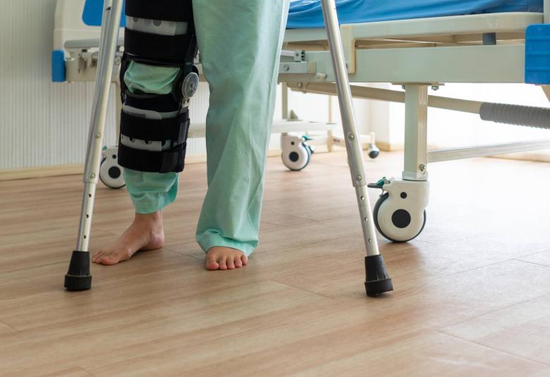 View from the thighs down of a patient wearing scrubs and a leg brace. The patient uses crutches to stand by a hospital bed.