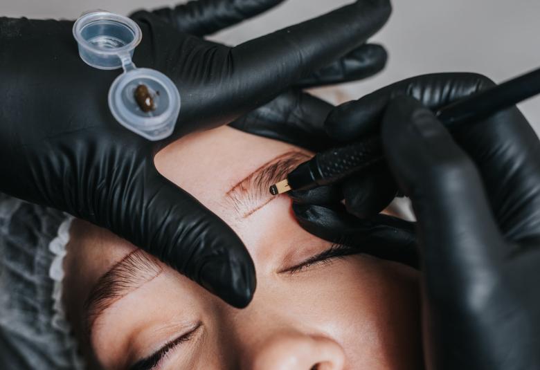 The gloved hands of a cosmetologist performing a microblading procedures along the eyebrows of a patient.