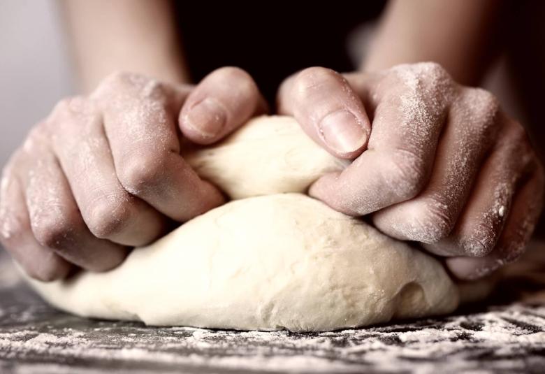 An extreme close-up view shows two flour-covered hands kneading dough on a flour-covered dark surface.