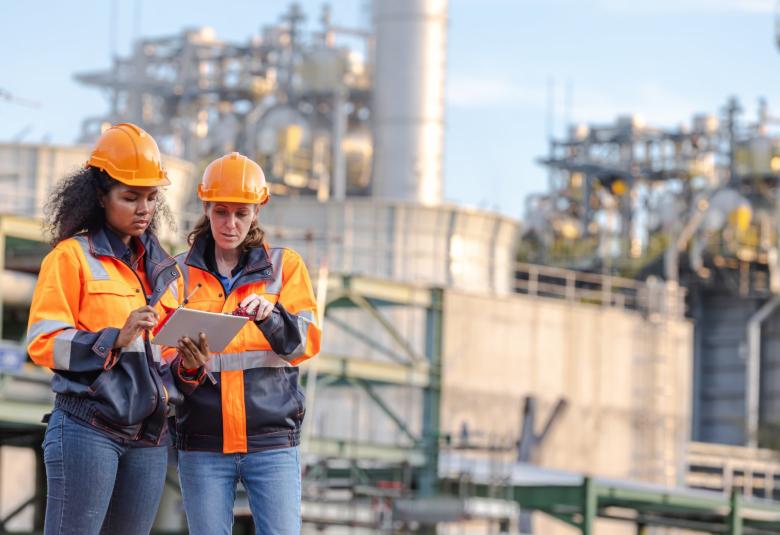 Two women wearing orange safety jackets and orange hard hats look at a tablet. Large pipelines are in the background.