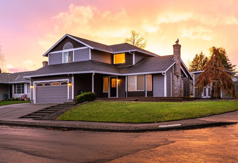 A well-maintained two-story house with a garage and walkway in a neighborhood. The sun is setting behind the house.