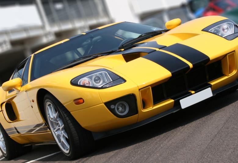 A yellow sports car is parked outside on a sunny day. There are black racing stripes on the top and side of the vehicle.