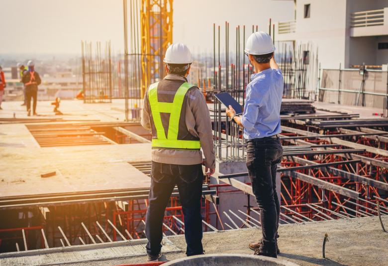 Two people in hard hats standing together while looking at an active construction site. One is holding a tablet and pointing.
