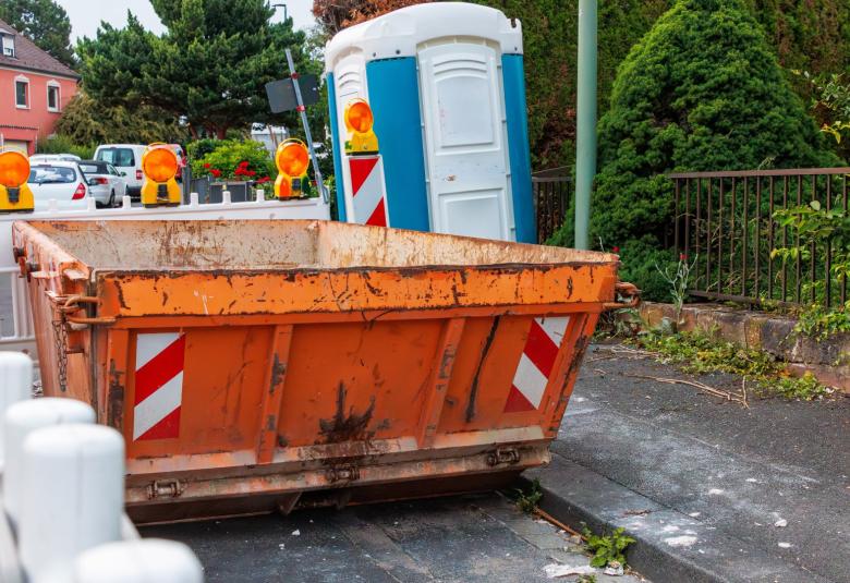 A large, metal dumpster that is orange in color is sitting on the side of a road that is under construction.