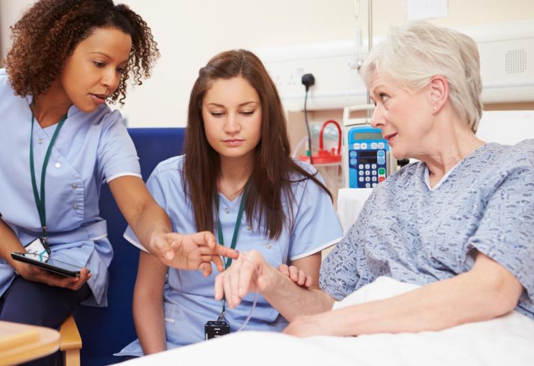 A senior nurse instructs a student nurse at a patient's bedside inside a hospital room during a clinical training visit.