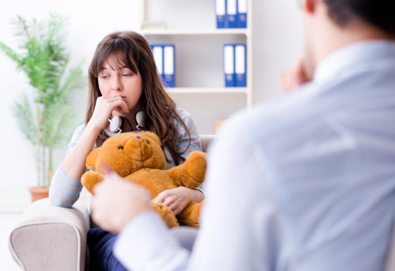 A young girl sitting with a teddy bear in front of a talk therapist. She has her hand on her mouth and she's looking down.