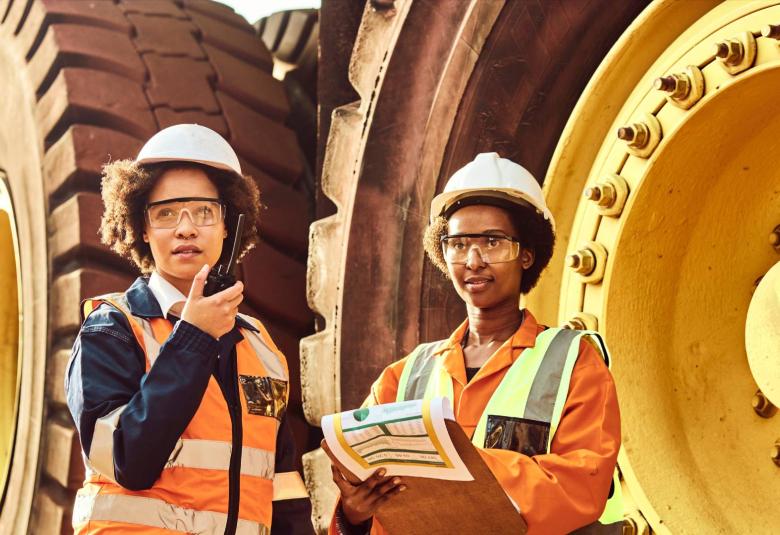  Two female mine workers wearing hi-vis vests and hard hats, surveying mining equipment on a site and writing on a clipboard.