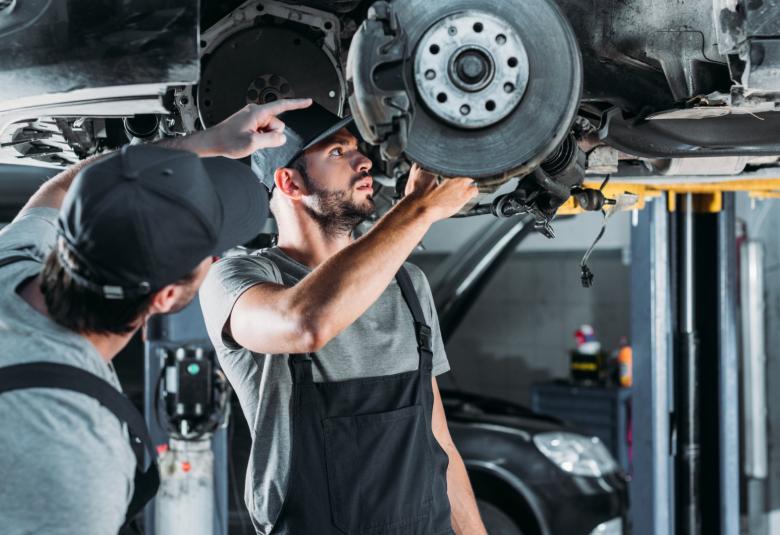 Two mechanics working on the underside of a car in an auto repair shop. The car is raised above the mechanics.