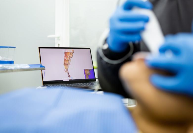 A dentist in blue gloves uses an intraoral scanner on a patient, with a laptop displaying the digital scan in the background.