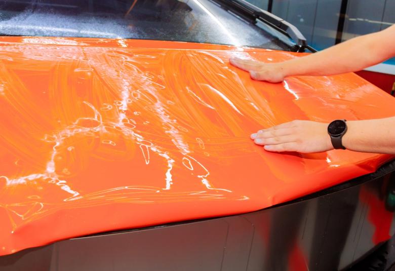 A person with a black watch on their left arm uses both hands to smooth out an orange vinyl wrap over the hood of a car.