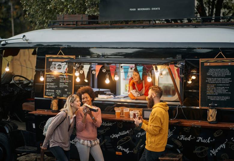 A group of three individuals share food in front of a stylish, modern-looking food truck. A worker smiles at them from inside.