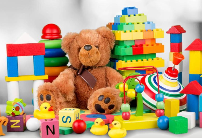 A pile of colorful toys on a plain white desk, including a stuffed bear, building blocks, alphabet blocks, and rubber ducks.