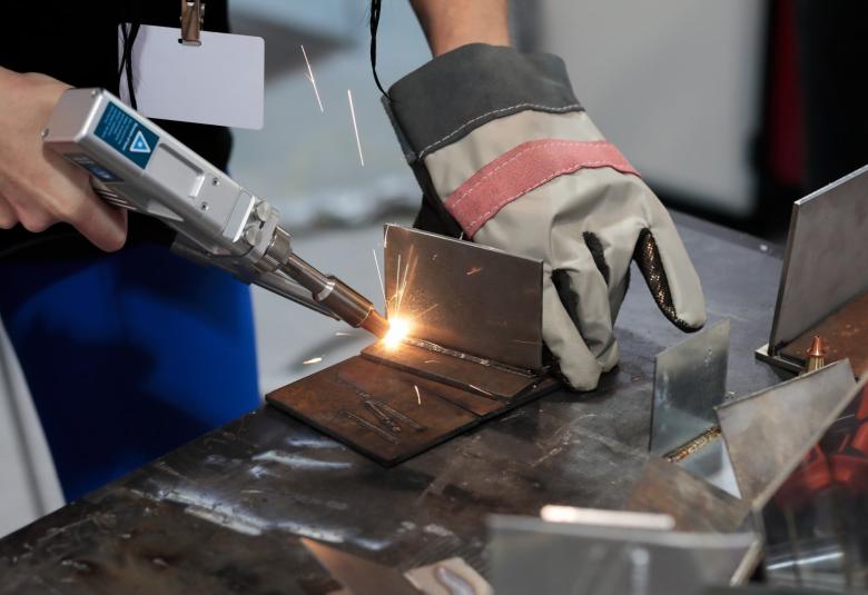 A person standing at a workstation using a portable welder to bind together pieces of metal. They're have gloves on.