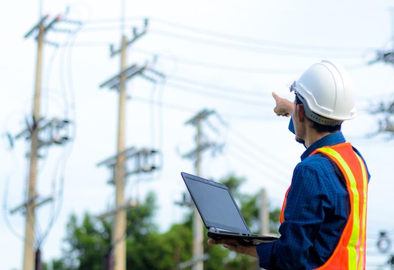 A utility worker wearing PPE hat and vest and holding a laptop computer points at power lines in the distance.