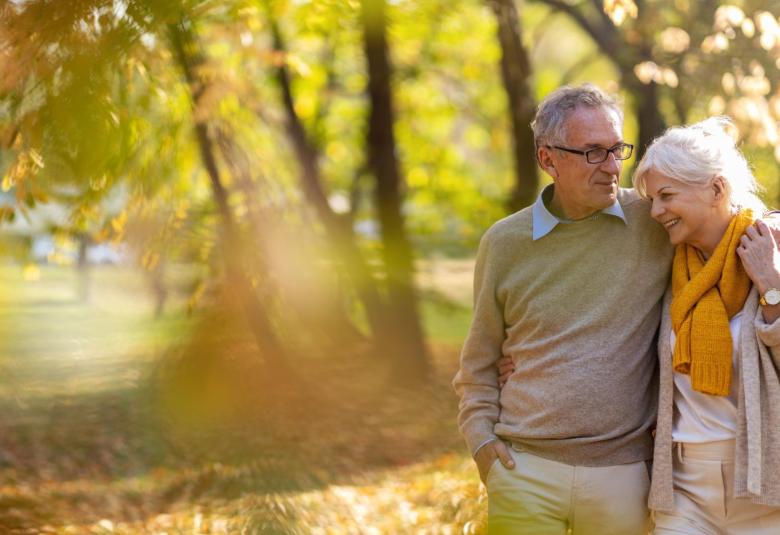 Two seniors walking through a park as leaves fall in the autumn season, wearing sweaters and scarves to stay warm.