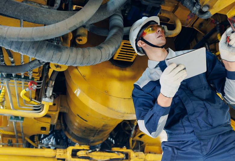 A man in a hard hat and protective glasses standing underneath a large piece of equipment. He is inspecting it.