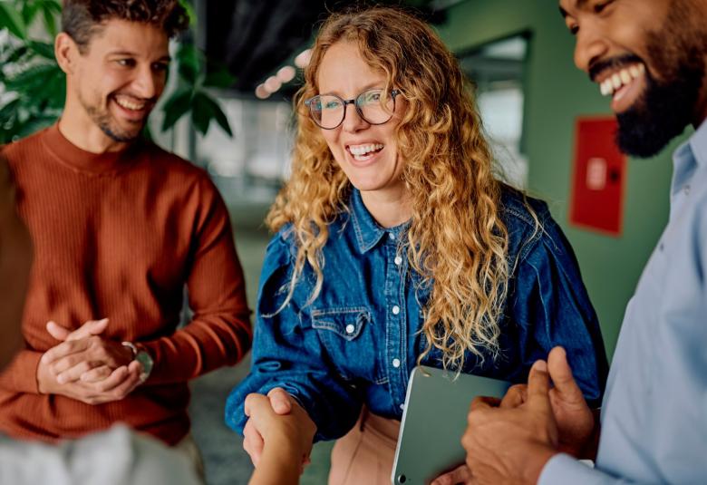 A group of employees stand together as the new female employee shakes the leaders hand with a smile on her face.