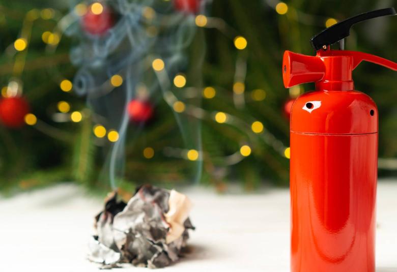 A fire extinguisher and a smoldering scrunched piece of paper are in the foreground with Christmas tree lights behind them.