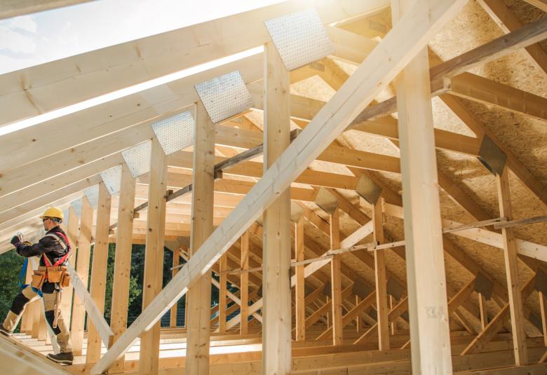 A worker securing lumber on a wooden roof frame at a residential construction site with safety gear beneath an open sky.