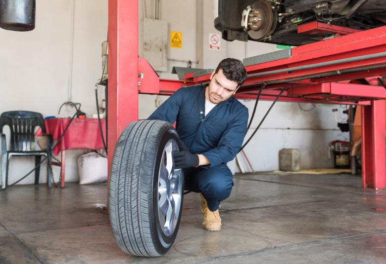 A mechanic in a blue jumpsuit changes a tire on a white car that's sitting on a red four-post auto lift.