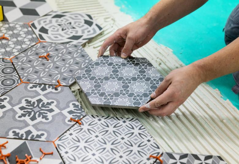 A man kneels down and lays different patterned, white and gray hexagon cement tiles on the floor of his home.