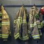 Three firefighter jackets hang alongside two red helmets on black hooks attached to a black cement wall.